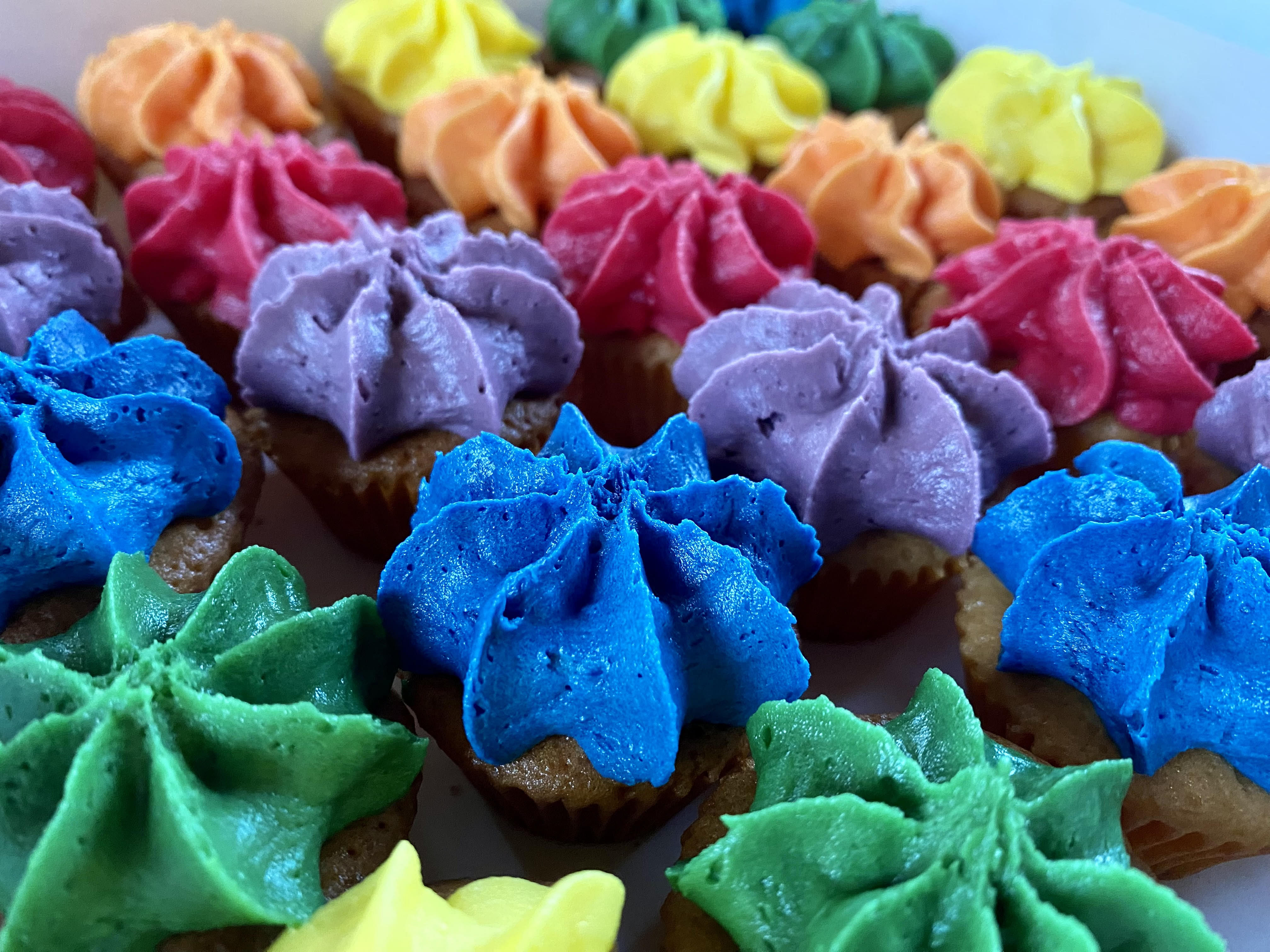 A close-up of rows of mini cupcakes with brightly colored, star-shaped frosting in the colors of the rainbow, including green, blue, purple, pink, orange, and yellow.